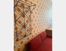 Innenraum mit einer langen Wand in orange-brauner, geometrisch-floraler Retro-Tapete und einem schwarzen Metall-Garderobenständer mit Haken. Im Vordergrund steht ein rotes Polstersofa, am rechten Rand ist ein helles Holzregal zu sehen.