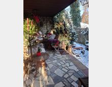 Überdachte Terrasse mit gefliestem Steinboden, Sitzmöbeln aus Rattan und Holztischen, dekoriert mit Pflanzen und roten Ornamenten. Im Hintergrund ein verschneiter Garten mit Holzzaun und immergrünen Bäumen.