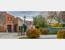 Straßenszene mit einer alten roten Backsteinfassade, einem modernen Tor und angrenzenden Gebäuden aus verputztem Stein. Im Vordergrund sind Hecken und ein Gehweg, während im Hintergrund Bäume und ein bemalter Zaun zu sehen sind.