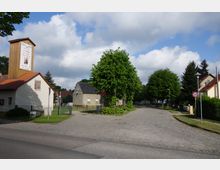 Straßenszene in einer ländlichen Umgebung mit gepflasterten Wegen, einem kleinen weißen Gebäude mit Holzdach und Werbeplakat, umgeben von Bäumen und Wohnhäusern. Ein "30"-Verkehrsschild steht am Straßenrand, und der Himmel ist teils bewölkt.