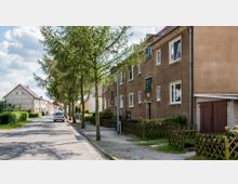 Straßenszene in einem Wohngebiet mit zweigeschossigen Häusern in schlichter Bauweise, braunen Fassaden und weißen Fenstern. Auf der linken Straßenseite stehen Bäume und geparkte Autos, umgeben von kleinen Vorgärten und Holzzäunen.