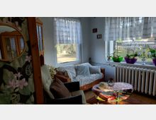 Gemütlicher Wohnbereich mit Rattansofa und Sitzmöbeln vor zwei großen Fenstern mit halbtransparenten Vorhängen. Auf der Fensterbank stehen Pflanzen in farbigen Schalen über einem Heizkörper; davor ein runder Couchtisch auf Teppich.