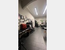 Innenraum eines modernen Tattoo-Studios mit schwarzen Liegen, Rollstühlen und Werkzeugwagen. Die Wände sind mit gerahmten Kunstwerken dekoriert, und im Hintergrund sind Pflanzen und weitere Einrichtungsgegenstände sichtbar.