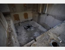 Blick in einen großen, leerstehenden Betonraum mit hohen Wänden und staubigem Boden; in der Mitte befindet sich eine rechteckige Öffnung mit Metallrahmen. An der Stirnseite sind zwei hohe Fenster mit Holzbrettern vernagelt, daneben sind raue Betonflächen und eine abgesenkte Nische erkennbar.