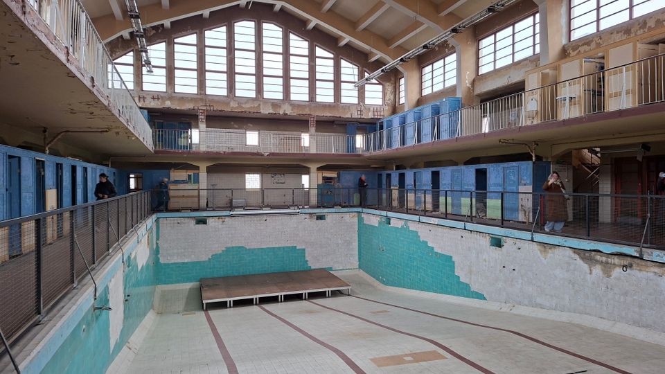 Ehemaliges Hallenbad mit leeren Becken und verblasster, türkisfarbener Fliesenverkleidung, umgeben von zwei Galerien mit blauen Umkleidekabinen und großen Fenstern, die im Obergeschoss viel Licht hereinlassen. Die Architektur weist sichtbare Alterungsspuren und abgenutzte Oberflächen auf.