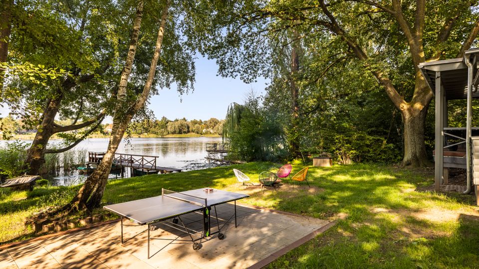 Ufernaher Garten am See mit schattenspendenden Bäumen, Steg am Wasser und Rasenfläche. Im Vordergrund stehen eine Tischtennisplatte auf einer Terrasse sowie mehrere bunte Gartenstühle; rechts ist ein überdachter Gebäudeteil zu sehen.