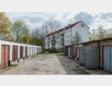 Eine Reihenanlage von Garagen mit bunt gestrichenen Holztoren befindet sich links und rechts eines unbefestigten Wegs. Im Hintergrund steht ein mehrstöckiges Wohnhaus mit weißer Fassade und roten Dachziegeln, umgeben von Bäumen.