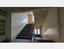 Treppenhaus in einem Gebäude mit gelb gestrichenen Wänden und einem Handlauf in Rot. Eine Treppe führt hinauf zu einem Fenster, das Tageslicht hineinlässt, und hinunter zu einer Tür mit Glaselementen.