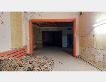 Ein leerer, renovierungsbedürftiger Innenraum mit abblätternder Wandfarbe, einer offenen Tür mit orangefarbenem Rahmen und einem dahinter liegenden dunklen Raum. Im Vordergrund liegt ein großer Haufen Holzlatten, und rechts ist eine helle Lampe an der Wand befestigt.