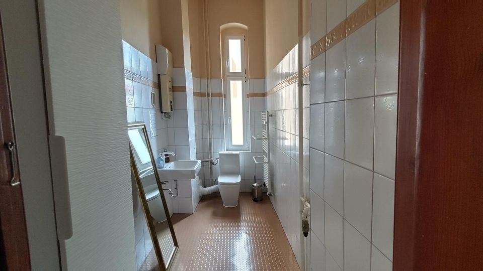 Schmales, längliches Badezimmer mit hohen Decken, weiß gefliesten Wänden mit Zierbordüre und braunem Bodenbelag. Am Ende des Raums stehen WC und ein hohes, schmales Fenster; seitlich sind ein kleines Waschbecken, ein Heizkörper und ein an die Wand gelehnter Spiegel zu sehen.