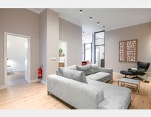 Modernes, offenes Wohnzimmer mit hellgrauem Ecksofa auf Holzfußboden, Glas-Couchtisch und schwarzem Relaxsessel neben einem Wandbild. Hohe Decken mit Einbauspots, breite Türöffnungen und eine Glastrennwand führen zu angrenzenden Räumen und einem hellen Fensterbereich.