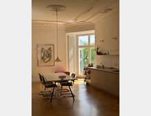 Wohnzimmer mit Essbereich in einem Altbau mit hohen Decken und verzierten Stuckelementen. Im Raum befinden sich ein Esstisch mit Stühlen, eine moderne Küchenzeile, große Fenster mit Blick ins Grüne und ein Kunstwerk an der Wand.