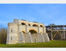 Historisches Bauwerk aus hellem Stein mit mehreren großen, bogenförmigen Durchgängen und befestigten Ecken, daneben eine erhöhte Stahlkonstruktion. Im Hintergrund sind Bäume und ein blauer Himmel zu sehen.