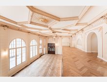Heller, leerer Innenraum mit großen bogenförmigen Sprossenfenstern, Fischgrätparkett und einer erhöhten Galerieebene. Die Decke ist reich mit Stuck und einem zentralen Sonnenmotiv verziert, dazu helle Wandpaneele und Rundbogenöffnungen.