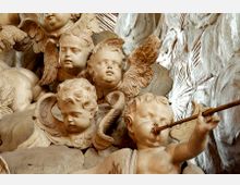 Barocke Skulpturengruppe mit mehreren Engelsköpfen und Putten, darunter ein musizierender Engel mit einer Flöte. Hintergrund aus hellen, organisch wirkenden Stein- oder Stuckelementen.