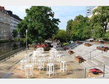 Freiluft-Veranstaltungsort an einem Flussufer mit weißen Stehtischen auf einer Terrasse, einer Sitztreppe mit bunten Kissen, großen grünen Bäumen und umliegenden modernen Gebäuden. Im Hintergrund befinden sich weitere Sitzmöglichkeiten sowie eine Bar.