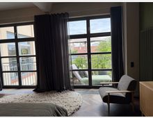 Modern bedroom with herringbone wood flooring, a round shag rug, and an upholstered chair beside floor-to-ceiling glass doors with dark curtains. The doors open to a balcony with outdoor seating and greenery, with neighboring buildings visible outside.