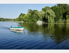 Ein See mit ruhigem Wasser umgeben von dichtem Grün und hängenden Weiden; im Vordergrund fährt ein kleines Motorboot, im Hintergrund ist ein weiteres Boot an einem kleinen Steg vertäut.