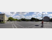 Weitläufige Kreuzung in einer Stadt mit markierten Radwegen, umgeben von Gebäuden im klassischen Stil und Bäumen. Links befindet sich ein geschwungenes Gebäude, rechts ein rundes Eckgebäude, und im Vordergrund fährt eine Person auf einem Fahrrad.