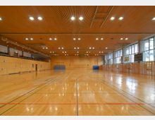 Große, helle Sporthalle mit Holzfußboden und markierten Spielfeldern, umgeben von holzvertäfelten Wänden und großen Fenstern, die Tageslicht hereinlassen. An der Decke befinden sich zahlreiche runde Lampen, an den Wänden sind Sportgeräte und eine Tribüne erkennbar.
