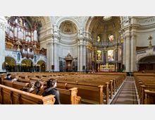 Innenraum einer großen Kirche mit reich verzierter Architektur, hohen Säulen, einer Kuppeldecke und kunstvollen Fenstern. Im Vordergrund stehen hölzerne Kirchenbänke, während links eine große Orgel und rechts der Altar mit goldenen Verzierungen zu sehen sind.