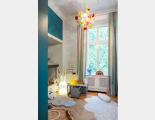 Kinderzimmer mit Holzfußboden, buntem Kronleuchter und großen Fenstern mit gestreiften Vorhängen. Im Raum befinden sich Spielzeug, ein kleiner Tisch mit Stühlen, leuchtende Tierlampen und Teppiche in Wolkenform.