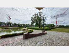 Außenansicht eines gepflasterten Geländes mit einem kleinen Teich im Vordergrund, einer symmetrischen Ziegelmauer und Grünanlagen. Im Hintergrund stehen moderne, rot-weiße Funktürme sowie mehrere Backsteingebäude, umgeben von Bäumen und Wiesen.