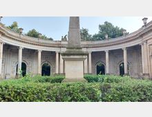 Halbrunde Kolonnaden mit dekorativen Säulen und Reliefverzierungen umgeben einen zentralen Obelisken aus Stein, vor dem dichte, gepflegte Sträucher angeordnet sind. Im Hintergrund sind Bäume und der offene Himmel sichtbar.