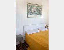 Ein schlicht eingerichtetes Schlafzimmer mit weiß gestrichenen Holzpaneelen an der Wand, einem Doppelbett mit gelber Bettdecke und weißen Kissen sowie einer an der Wand hängenden Weltkarte in einem türkisfarbenen Rahmen. Links neben dem Bett steht ein kleiner Tisch mit einer grauen Decke, und auf dem Boden liegt ein gestreifter Teppich.