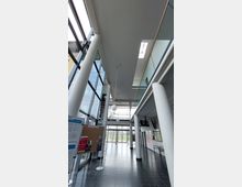 Innenansicht eines modernen Gebäudes mit hohen Decken, großen Glasflächen und weißen Säulen. Im Vordergrund sind Pinnwände mit Aushängen, während im Hintergrund Glastüren den Blick auf eine Rasenfläche freigeben.