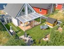 Luftaufnahme eines modernen Einfamilienhauses mit grau gedecktem Satteldach und großen Fenstern. Im Garten befindet sich eine überdachte Terrasse mit Holzboden, ein Grillbereich, ein kleines Holzsaunahaus in Fassform und Sitzmöbel sowie gepflegter Rasen mit dekorativen Steinwegen.