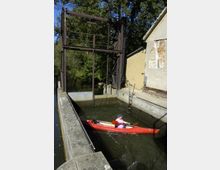 Eine Schleusenanlage mit einem roten Kajak, das von einer Person durch den schmalen Wasserweg gesteuert wird. Im Hintergrund sind ein metallisches Hubtor, ein Gebäude mit bröckelnder Fassade und Bäume zu sehen.