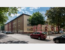 Wohnsiedlung mit mehrstöckigen Gebäuden in schlichtem Stil, teils beige, teils rosa gestrichen, mit Balkonen und Fenstern. Die Straße im Vordergrund zeigt parkende Autos, Fußgänger und Bäume, die teilweise Schatten spenden.