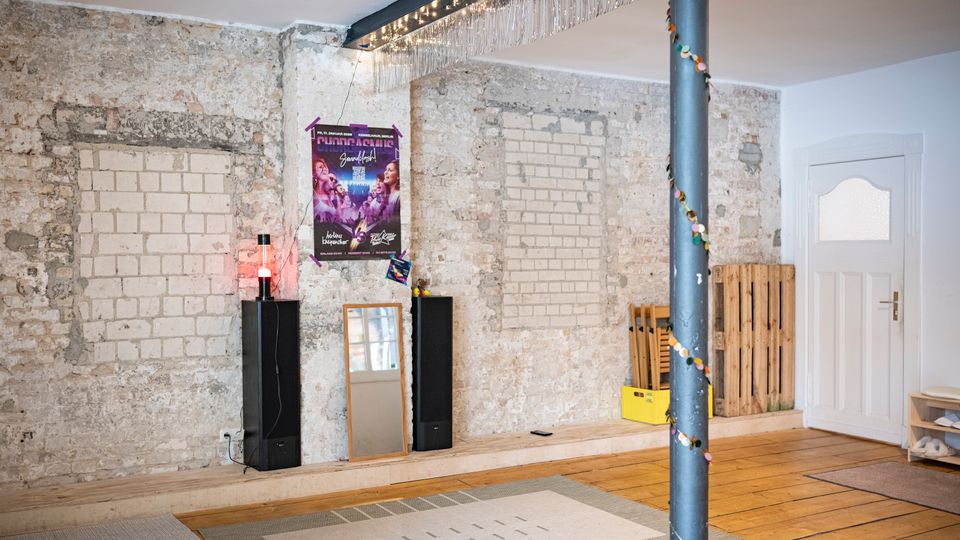 Innenraum mit freigelegter, rauer Ziegel- und Putzwand, Holzfußboden und einer dekorierten Stahlstütze. An der Wand hängen ein Poster und Lichterketten; davor stehen Lautsprecher, ein Spiegel sowie Holzpaletten neben einer weißen Tür.