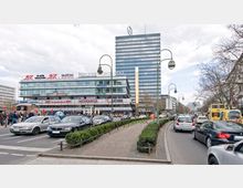 Straßenszene mit starkem Verkehr in einer Stadt, flankiert von modernen Gebäuden, darunter ein niedrigeres Gebäude mit Geschäften und Restaurants sowie ein hohes Bürogebäude im Hintergrund. In der Mitte der Straße befindet sich ein begrünter Mittelstreifen mit Lampenpfosten.