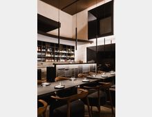 Ein stilvoll eingerichtetes Restaurant mit modernen schwarzen Tischen, Holzelementen und gedeckten Plätzen. Im Hintergrund befindet sich eine Bar mit dunklen Fronten und einer beleuchteten Rückwand, auf der Flaschen und Gläser stehen.