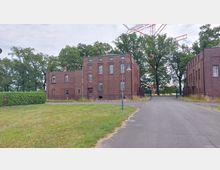 Backstein-Gebäude in historischer Industriearchitektur, mit quadratischen Fenstern und flachem Dach, umgeben von Bäumen und einer Grünfläche. Ein asphaltierter Weg führt zwischen den Gebäuden hindurch, dahinter ist ein Gittertor zu sehen.