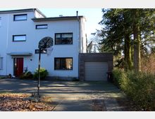 Zweistöckiges, modernes Wohnhaus mit weißer Fassade, großen rechteckigen Fenstern und einer roten Eingangstür. Ein Garagenanbau mit Holzverkleidung und grauem Tor befindet sich auf der rechten Seite, davor steht ein Basketballkorb, und die Umgebung ist von Bäumen und Büschen gesäumt.