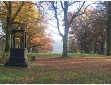 Ein herbstlicher Park mit einer Rasenfläche, umgeben von Bäumen mit gelbem und braunem Laub. Im Vordergrund steht ein dunkelgrünes, rechteckiges Denkmal mit Säulen, und der Boden ist mit Herbstblättern bedeckt.