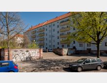 Mehrstöckiges Wohngebäude mit weißer Fassade und roten Ziegeldächern, umgeben von Bäumen. Im Vordergrund eine Mauer mit Graffiti, davor geparkte Autos und ein gepflasterter Platz mit Treppen.