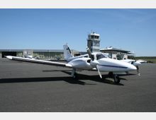 Ein kleines zweimotoriges Flugzeug steht auf dem Vorfeld eines kleinen Flughafens. Im Hintergrund sind ein Kontrollturm mit der Aufschrift "EDAZ" sowie Hangars und ein Gebäude mit einer Terrasse sichtbar, umgeben von einer flachen Landschaft und klarem Himmel.