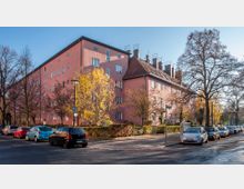 Eckgebäude eines mehrstöckigen Wohnkomplexes mit rosa Fassade und schrägem Ziegeldach, umgeben von kahlen und herbstlichen Bäumen. Parkende Autos säumen die Straße, die an der Kreuzung mit einem Straßenschild endet.