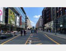 Straßenszene in einer Einkaufsstraße mit modernen Gebäuden aus Glas und Stahl auf beiden Seiten, darunter Geschäfte wie Galeries Lafayette und Karl Lagerfeld. Im Vordergrund sind gelbe Markierungen auf der Fahrbahn, Fußgänger, Fahrräder und ein kleines Beet sichtbar, bei blauem Himmel und Sonnenschein.