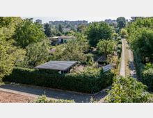 Blick auf eine Kleingartenanlage mit dicht bepflanzten Parzellen, Hecken und einem kleinen Gartenhaus mit Wellblechdach im Vordergrund. Im Hintergrund sind Wohnhäuser, Bäume und ein unbefestigter Weg zu sehen.