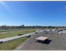 Freifläche mit Skatepark und Sitzgelegenheiten, umgeben von einer großen Wiese und vereinzelten Gebäuden im Hintergrund. Klare, blaue Himmelsszenerie mit asphaltierten Wegen, Rampen und einem bemalten Betonblock im Vordergrund.
