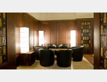 Ein Sitzbereich mit fünf schwarzen Ledersesseln um einen niedrigen Holztisch, umgeben von dunkelbraunen Holzvertäfelungen und dekorativen Glaspaneelen. Im Hintergrund sind hohe Leuchten mit hellem, diffusem Licht zu sehen.