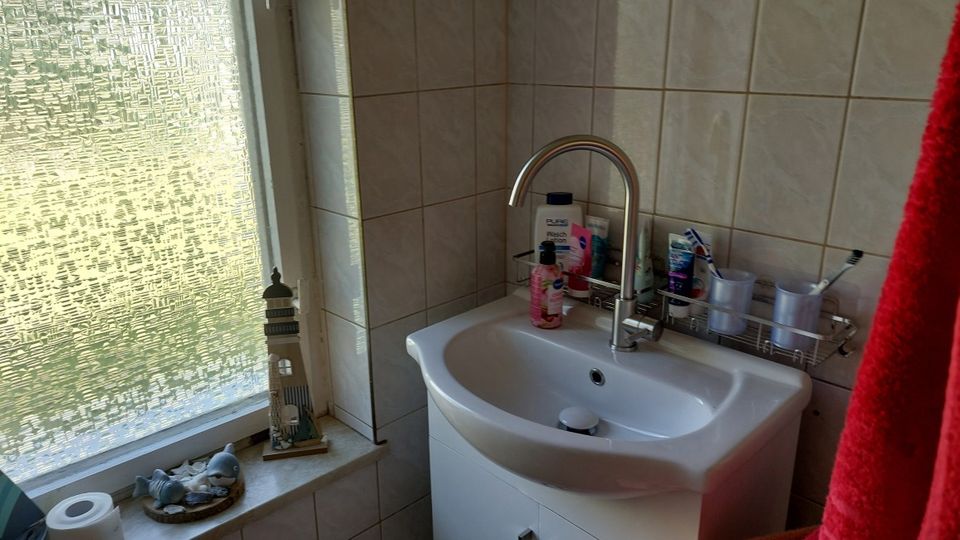 Waschbecken in einem gefliesten Badezimmer mit hohem Wasserhahn und Ablage mit Pflegeartikeln. Links befindet sich ein Fenster mit strukturierter Milchglasscheibe und Fensterbank mit Dekoration.