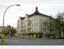 Ecke eines vierstöckigen Wohn- und Geschäftshauses im Gründerzeitstil mit heller Fassade, mehreren Balkonen und einem markanten Dachaufsatz. Im Erdgeschoss befindet sich ein Restaurant mit dem Schriftzug „Hoppegarten“, die Straße davor ist leer und von Bäumen sowie Straßenschildern gesäumt.