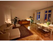 Gemütlicher Wohn- und Essbereich mit hellem Ecksofa, Teppich und Couchtisch sowie einem Esstisch mit Stühlen unter einer Hängelampe. Holzparkett im Fischgrätmuster, Sideboard mit Büchern, große Zimmerpflanzen und hohe Fenster mit Blick nach draußen.