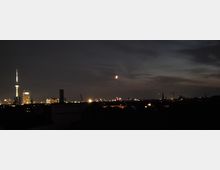 Nächtliche Stadtansicht mit einem beleuchteten Fernsehturm links im Bild und weiteren erleuchteten Gebäuden im Hintergrund, vor einem dunklen Himmel mit vereinzelten Wolken und einem leuchtenden Halbmond.